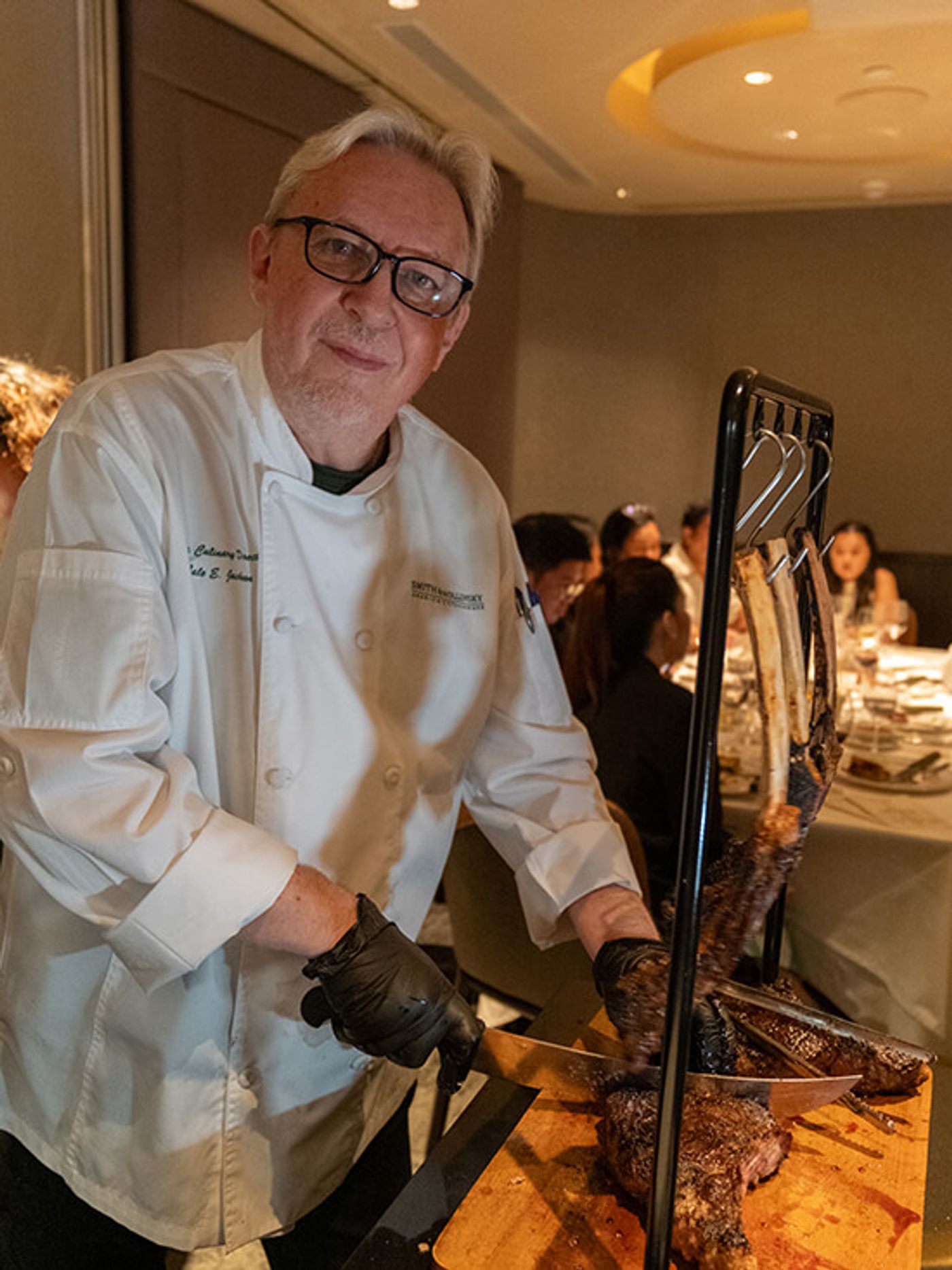 Chef Cale Jackson in chef whites and black gloves carving a Tomahawk steak beside seated diners