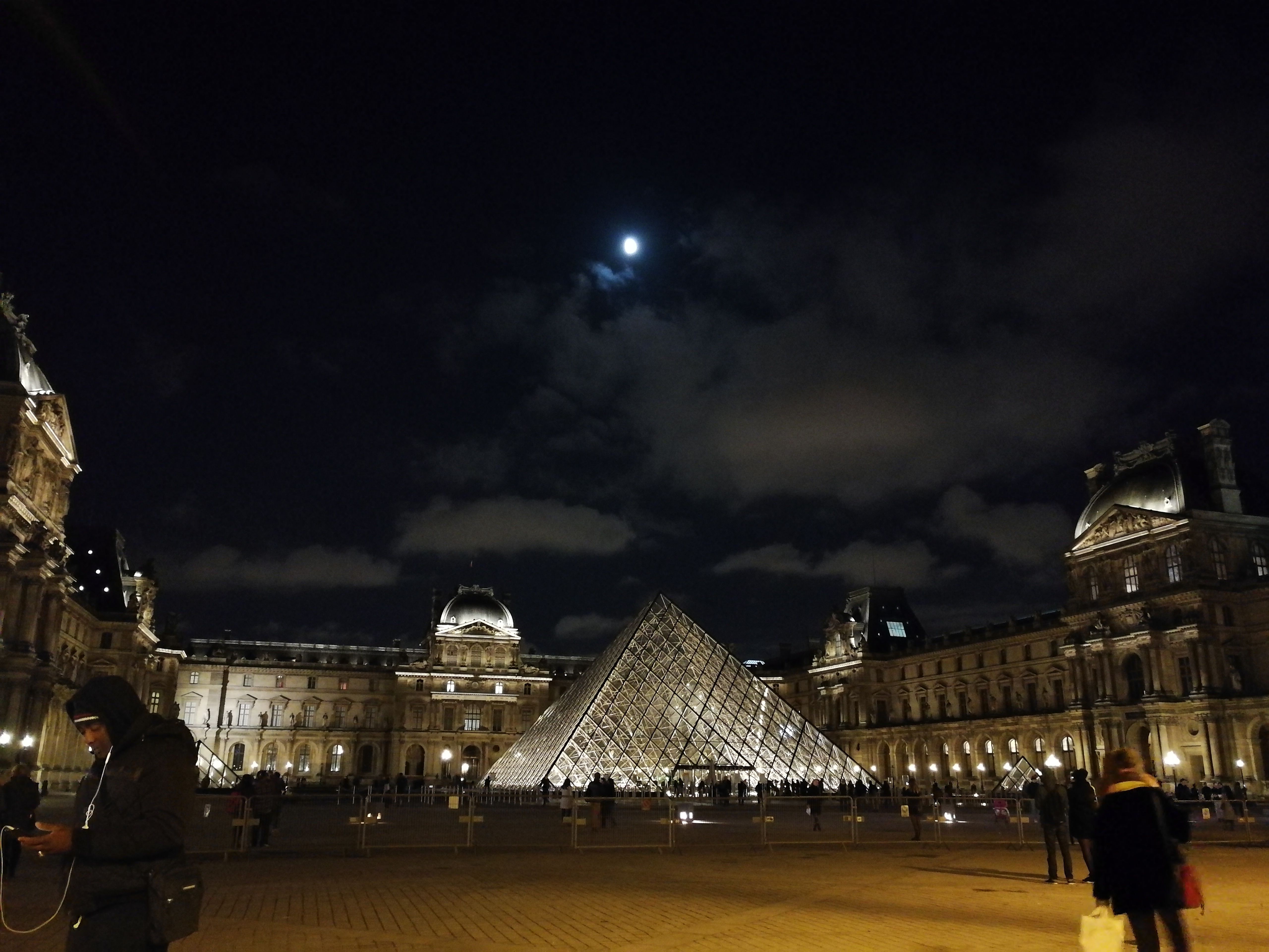 The Louvre’s glass pyramid glowing at night beneath a moonlit sky, crowds crossing the dark courtyard