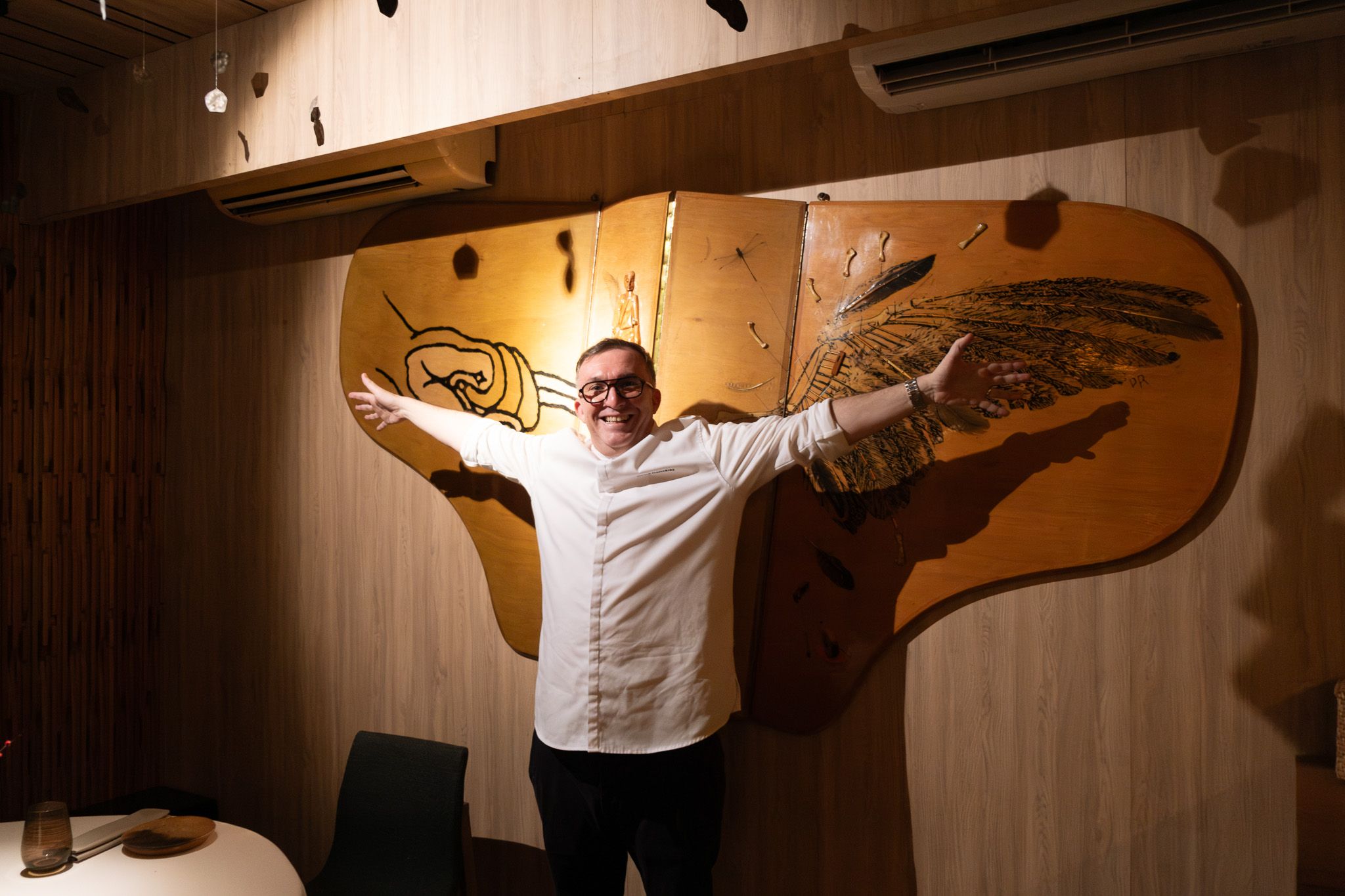 Chef Chele Gonzalez in whites spreads his arms wide before a winged wooden relief at Gallery by Chele