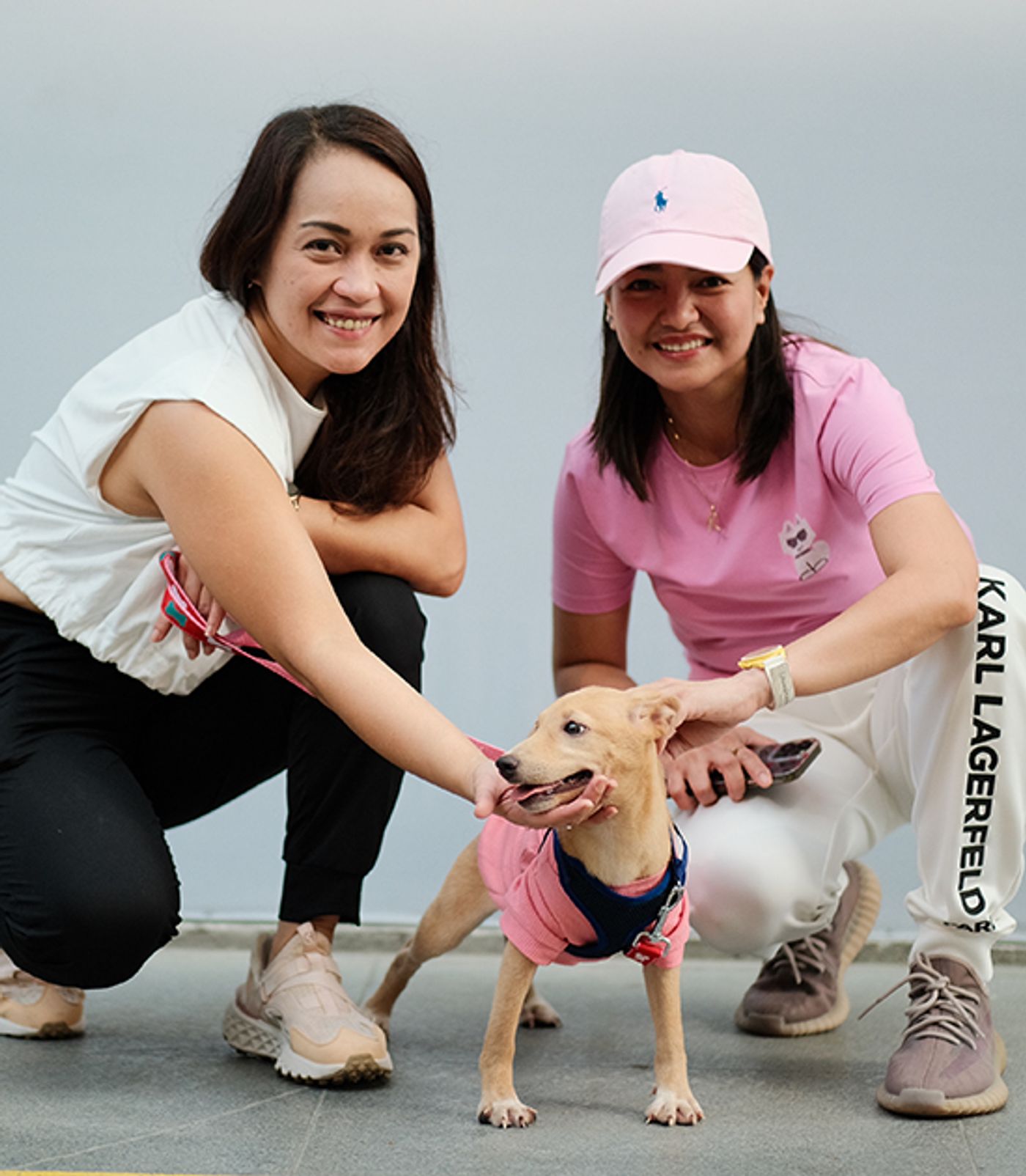 RC Tiongson and Jeremei Raagas crouch on either side of Olive, a small tan dog in a pink shirt, on a tiled floor