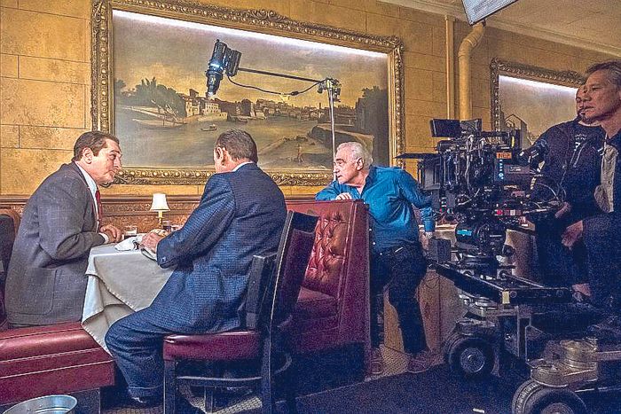 Martin Scorsese in a blue shirt directs a restaurant scene beside the camera as two actors sit at a table