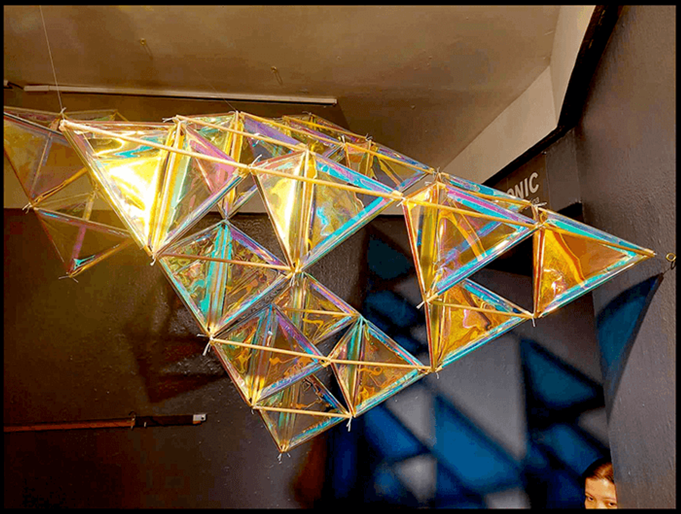 A tetrahedral kite of dichroic film glints gold, violet and teal, suspended from the ceiling at The Crucible gallery