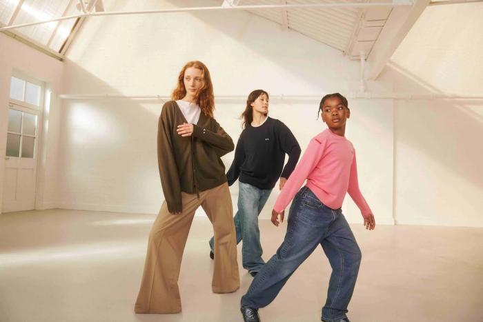 Three models mid-stride in a white studio wearing Uniqlo x Anya Hindmarch knitwear: an olive cardigan over a white tee with wide camel trousers, a navy crew-neck sweater with small embroidered eyes and loose jeans, and a bright pink sweater with wide jeans