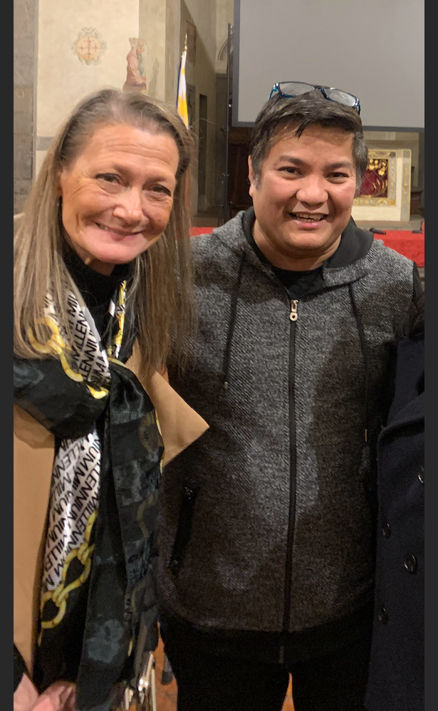 A woman with long grey hair in a patterned scarf and a smiling man in a grey hoodie, glasses perched on his head, pose inside a Venice church