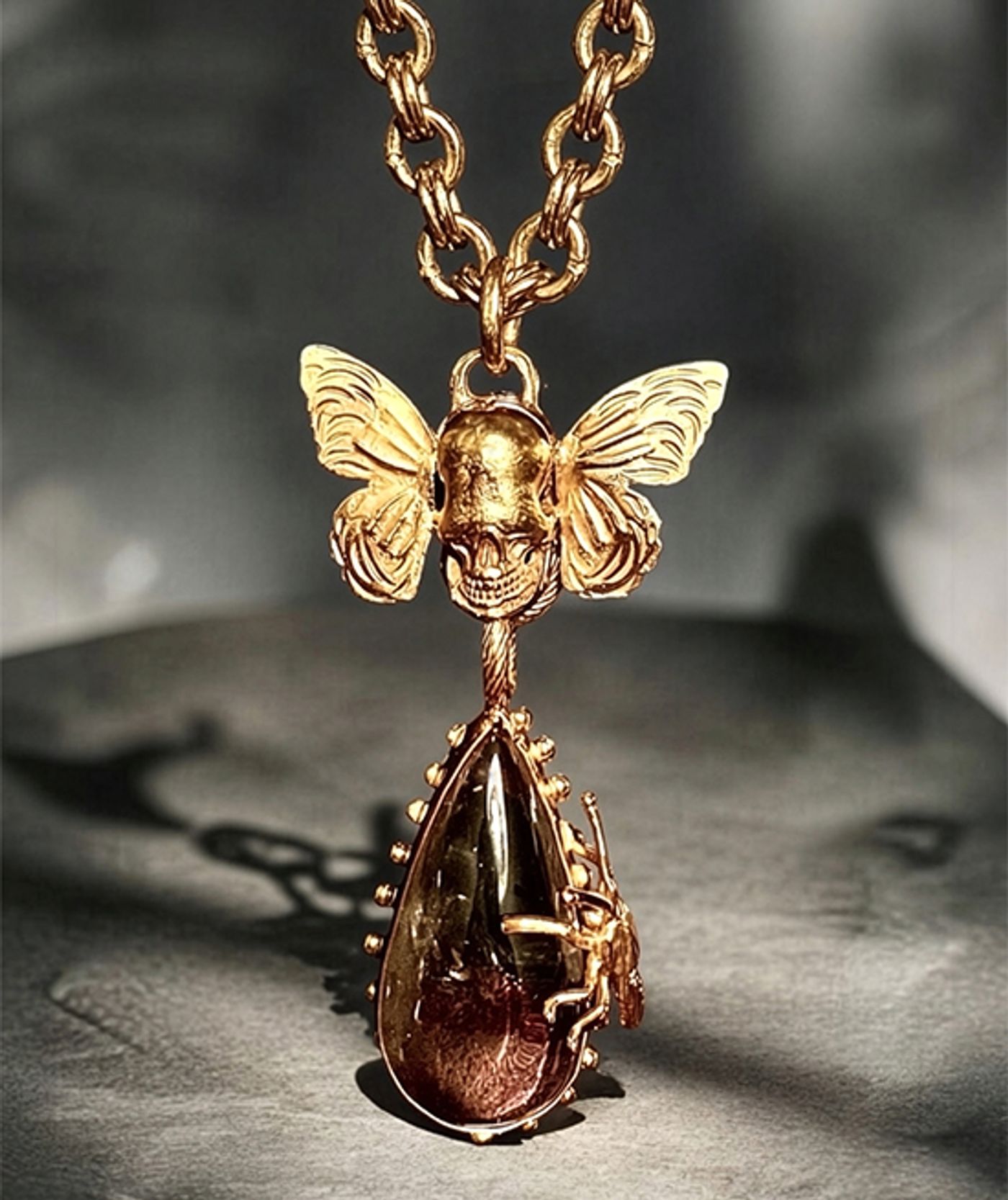 Gold pendant by Michelline Syjuco of a butterfly-winged skull above a teardrop quartz crystal on a heavy gold chain