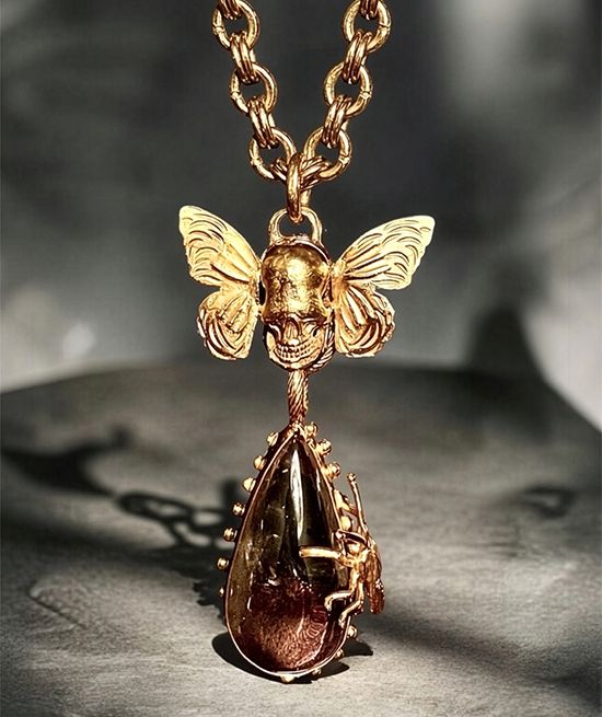Gold pendant by Michelline Syjuco of a butterfly-winged skull above a teardrop quartz crystal on a heavy gold chain