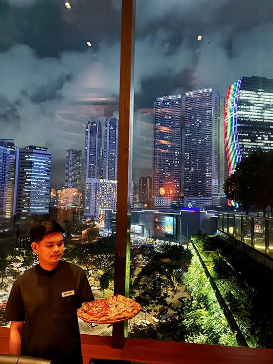 A server holds a pizza before a floor-to-ceiling window overlooking the illuminated Ortigas skyline at night