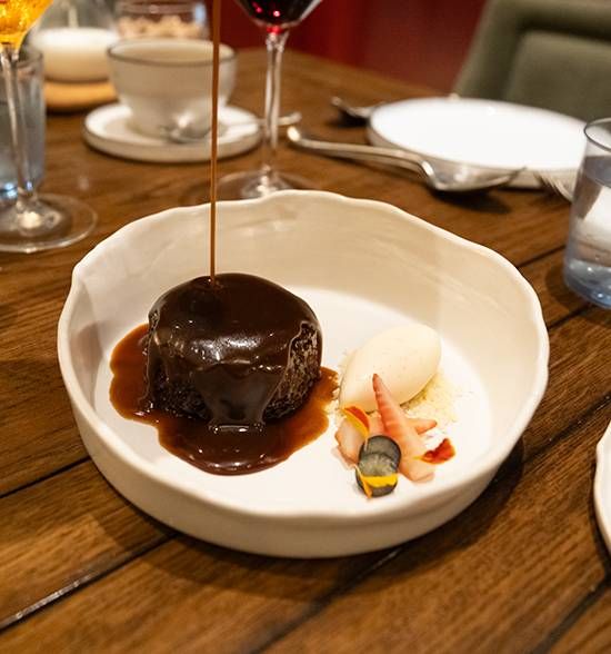 Warm toffee sauce being poured over a dark sticky toffee pudding beside a scoop of ice cream