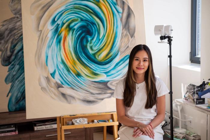 Maria Angelica Tan seated in her studio beside a large swirling blue and gold painting