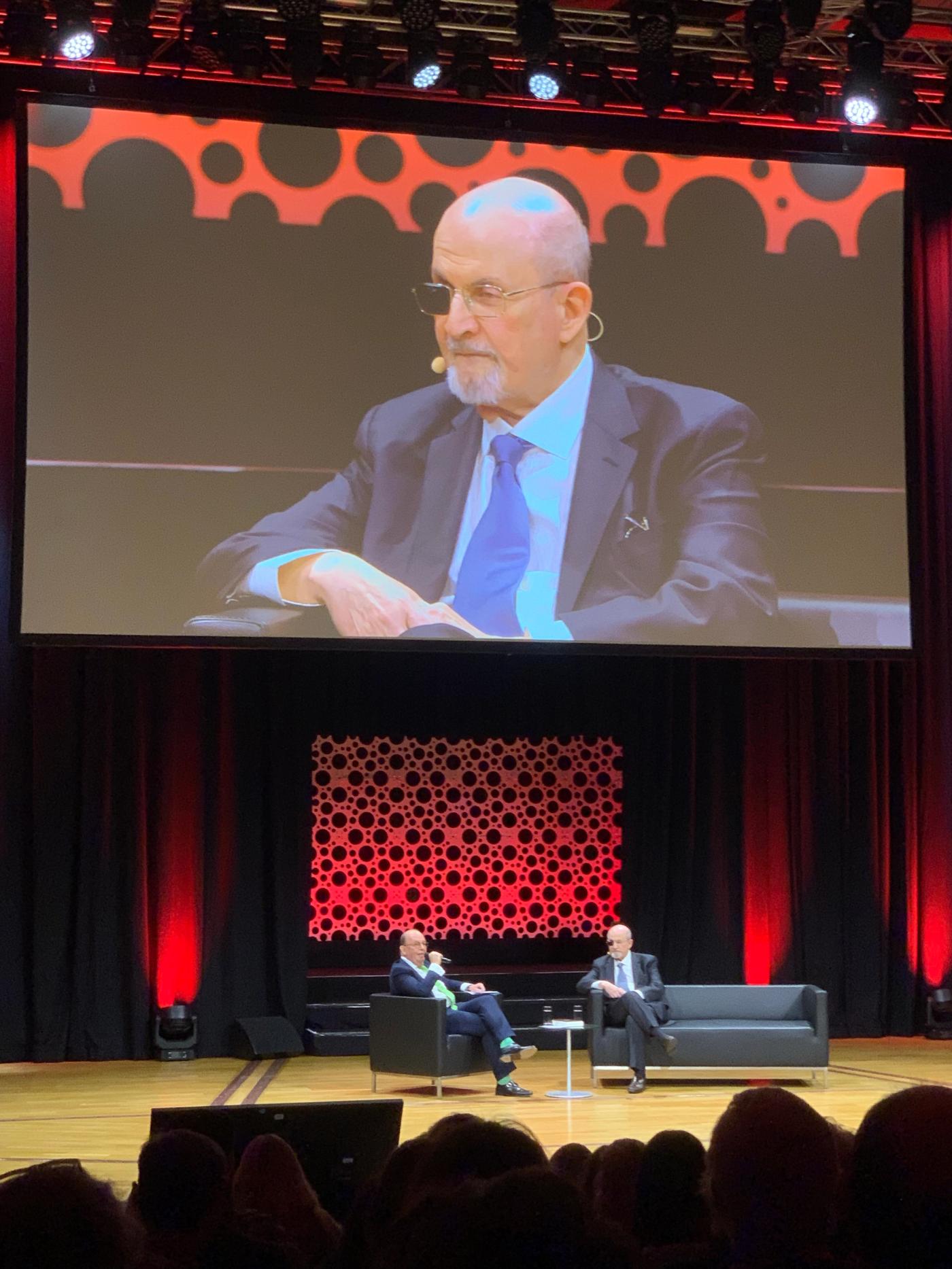 Salman Rushdie, in a dark suit and blue tie, speaks onstage at the Frankfurt Book Fair, magnified on the large screen above the red-lit stage before the audience