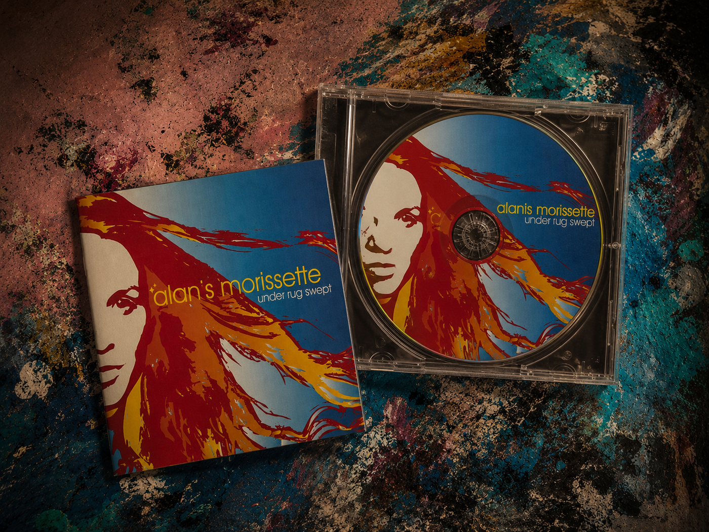 An Under Rug Swept CD tilted open beside its sleeve, Alanis Morissette’s flame-haired red-and-blue portrait doubled on disc and cover, on a paint-smeared tabletop
