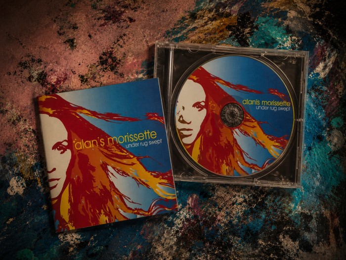 An Under Rug Swept CD tilted open beside its sleeve, Alanis Morissette’s flame-haired red-and-blue portrait doubled on disc and cover, on a paint-smeared tabletop