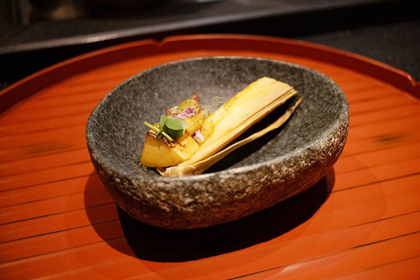 Sauteed Japanese bamboo shoot garnished with a leaf and blossom in a rough stone bowl on a red tray