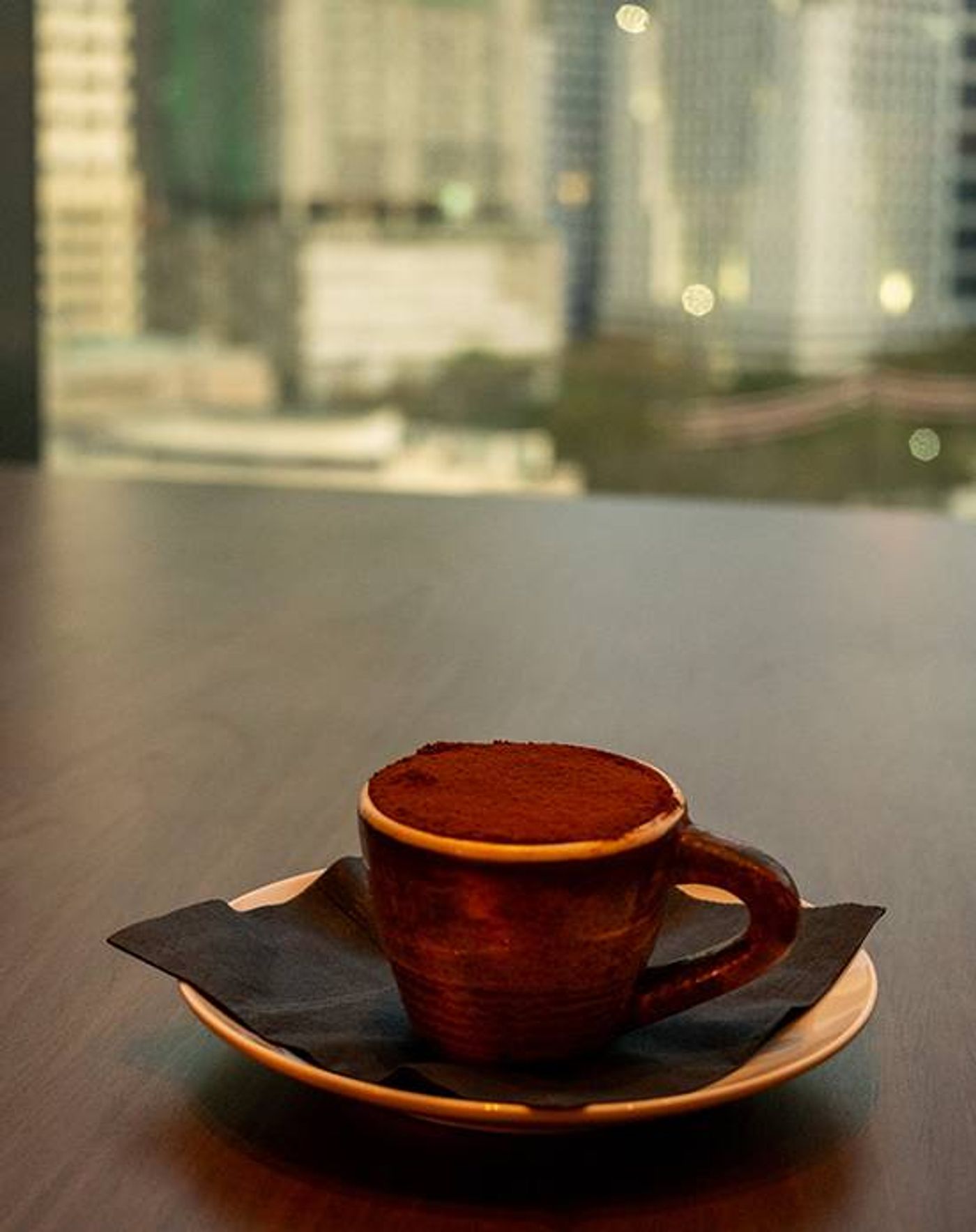 Cocoa-dusted tiramisu in a rustic ceramic cup on a dark saucer against Osteria Antica’s high-rise window view
