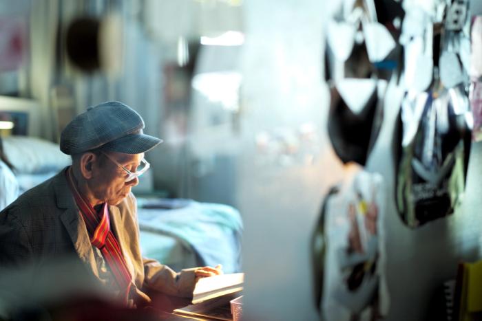 David Medalla in a grey flat cap, seen in profile in warm light, looking down at a book in a cluttered room