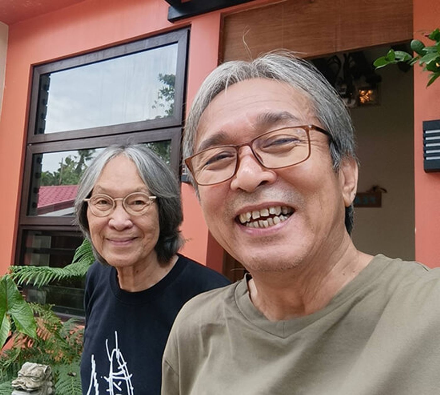 Selfie of a smiling man in glasses with a gray-haired companion outside an orange building