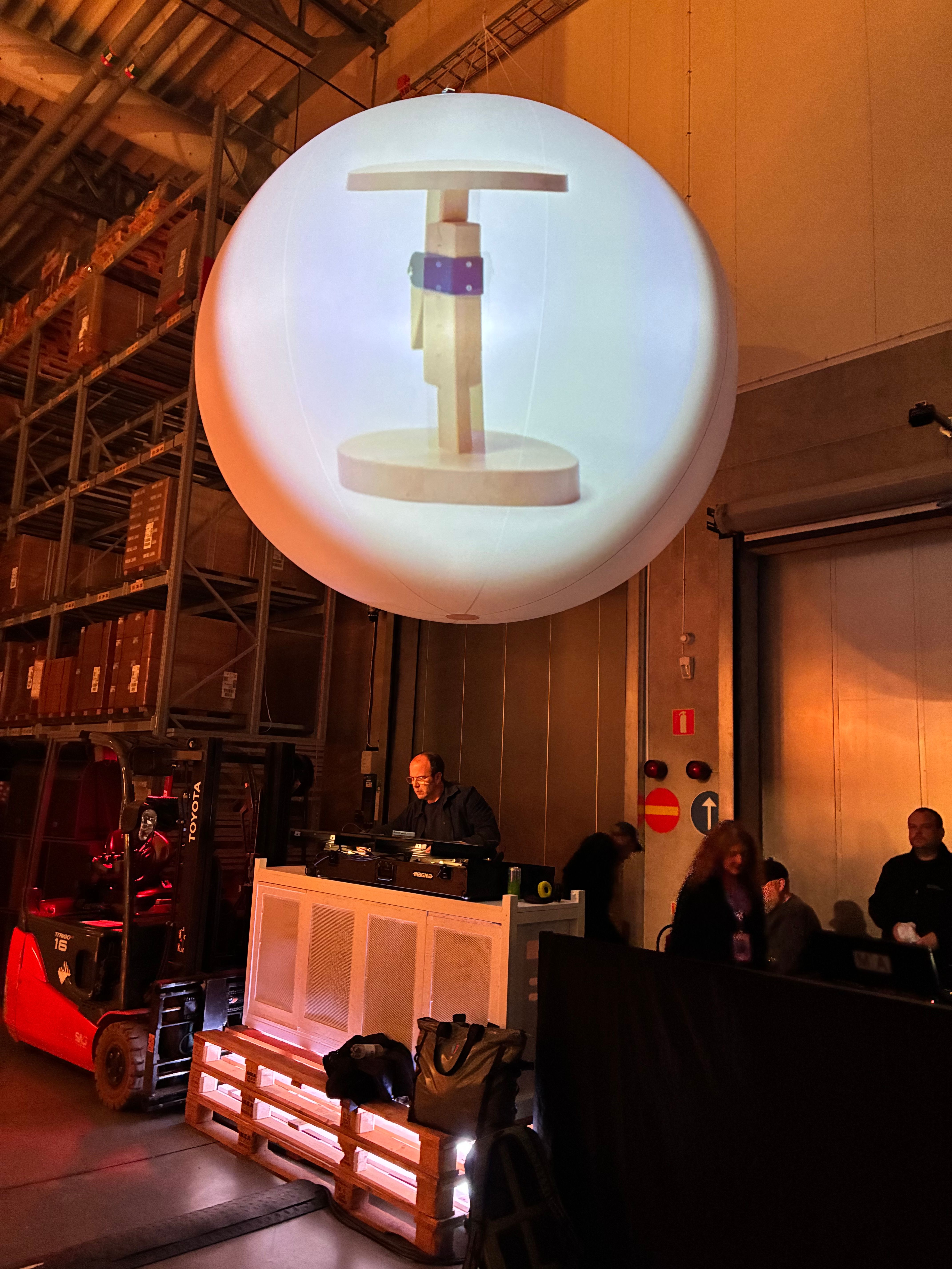Giant inflatable sphere projecting an IKEA PS 2026 valet stand above a DJ booth built on pallets, with a red forklift in the Älmhult warehouse