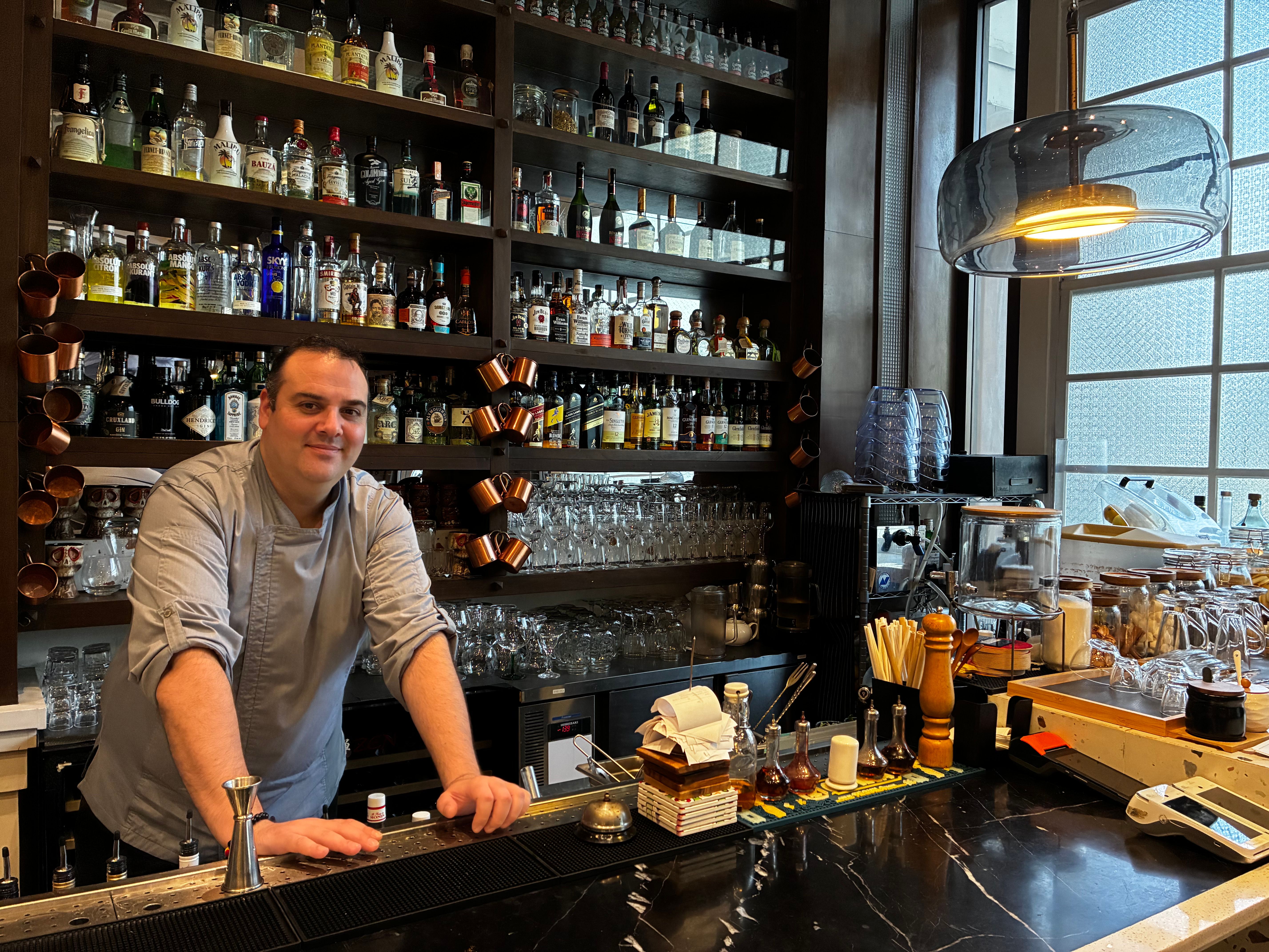 A chef leans on the black marble bar of a Bistronomia Group restaurant, backed by shelves of liquor bottles and copper mugs