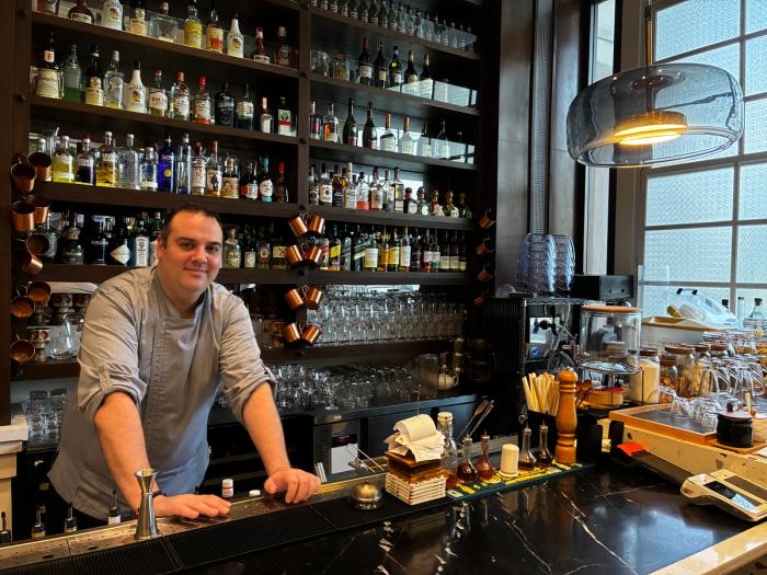 A chef leans on the black marble bar of a Bistronomia Group restaurant, backed by shelves of liquor bottles and copper mugs