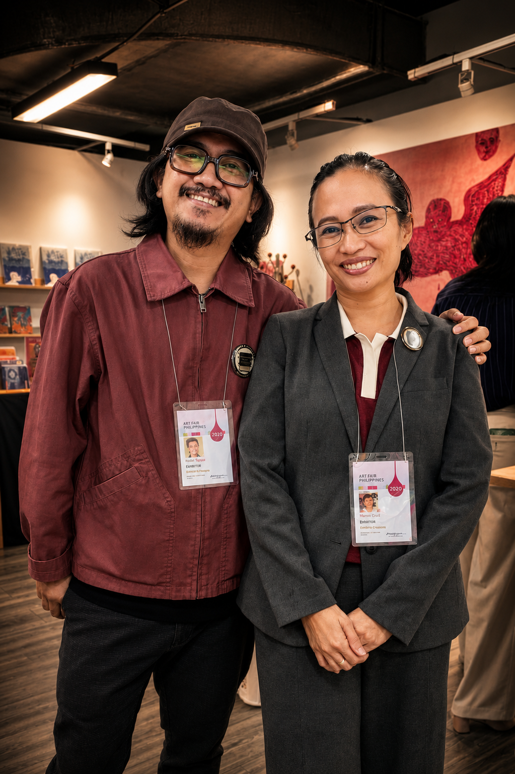 Rodel Tapaya in a cap and Marina Cruz in a grey suit, both in Art Fair Philippines 2020 exhibitor badges, smiling side by side in a gallery booth