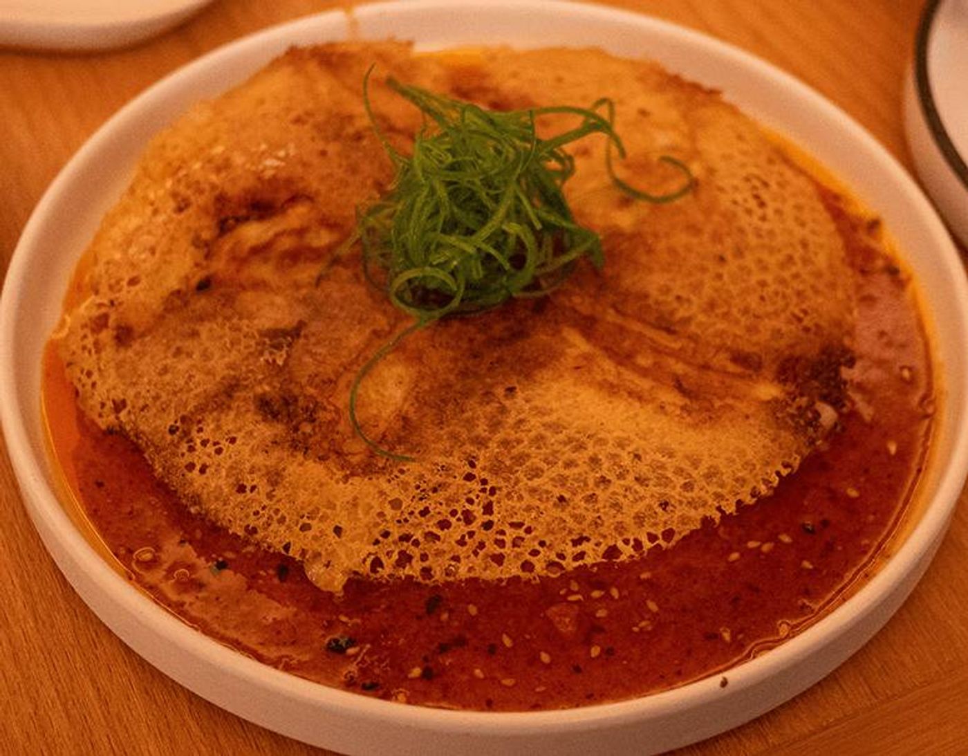 Spicy Hanetsuki Gyoza hidden under a lacy golden crepe wing, ringed by red chili oil and scallions