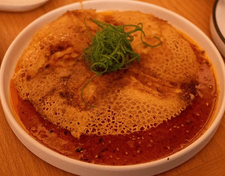 Spicy Hanetsuki Gyoza hidden under a lacy golden crepe wing, ringed by red chili oil and scallions