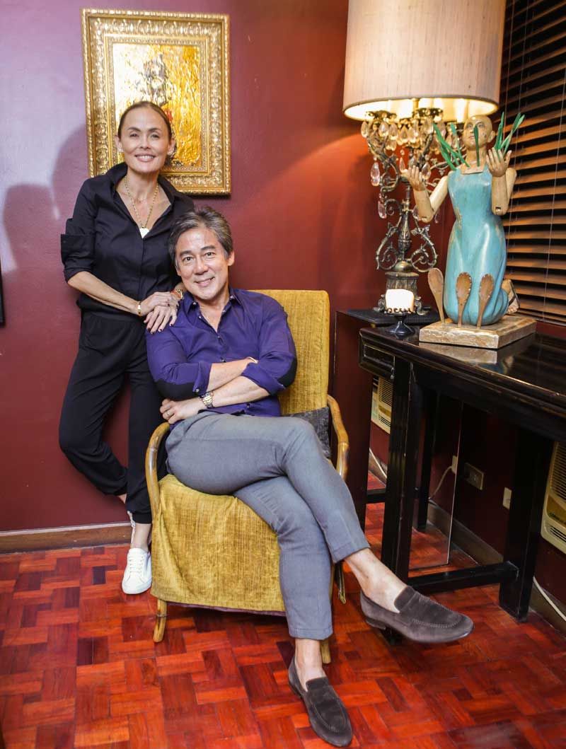 Joanna in black stands with a hand on Raul’s shoulder as he sits in a mustard armchair beside a carved blue figure lamp