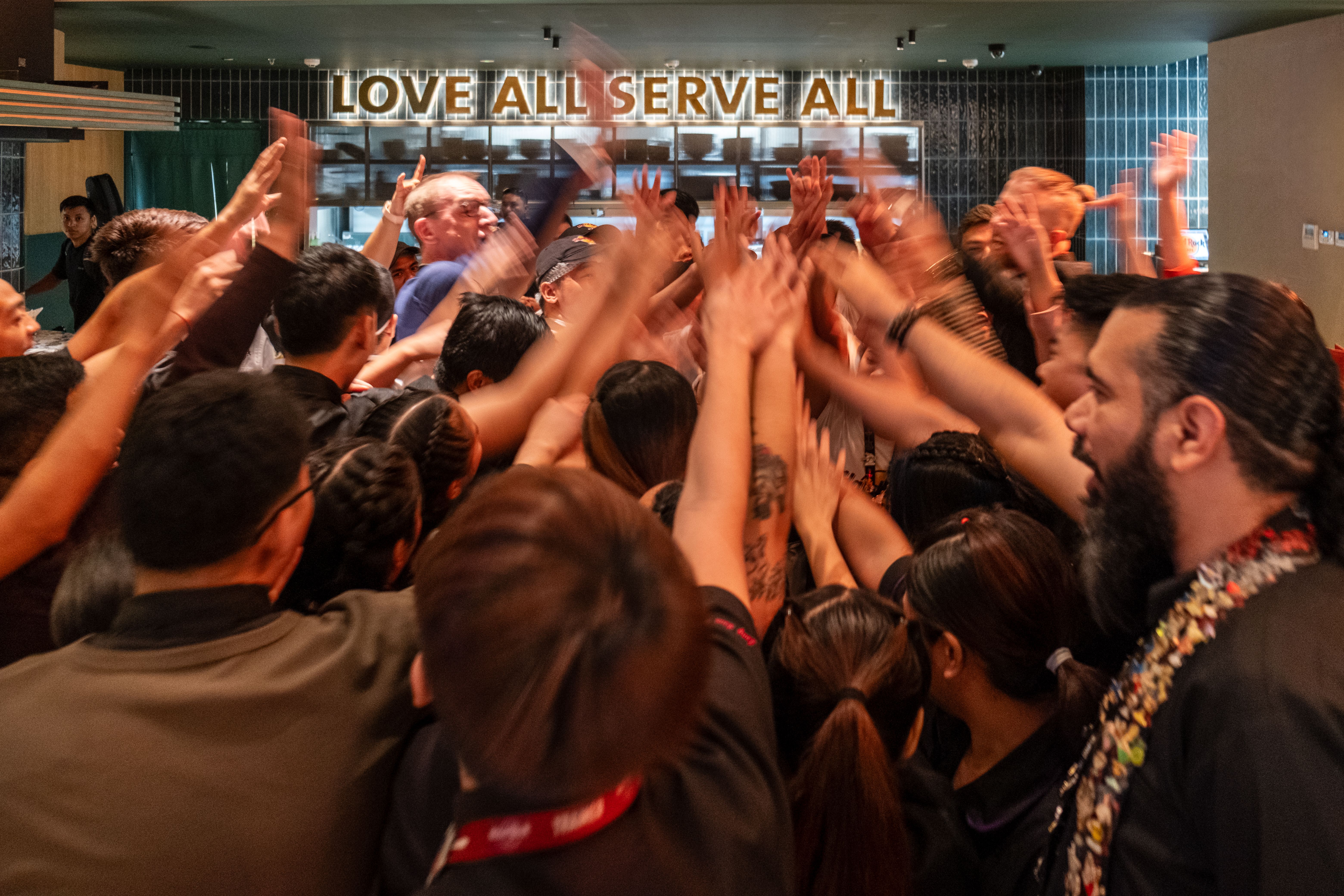 Hard Rock Cafe staff raise joined hands in a huddle beneath a Love All Serve All sign
