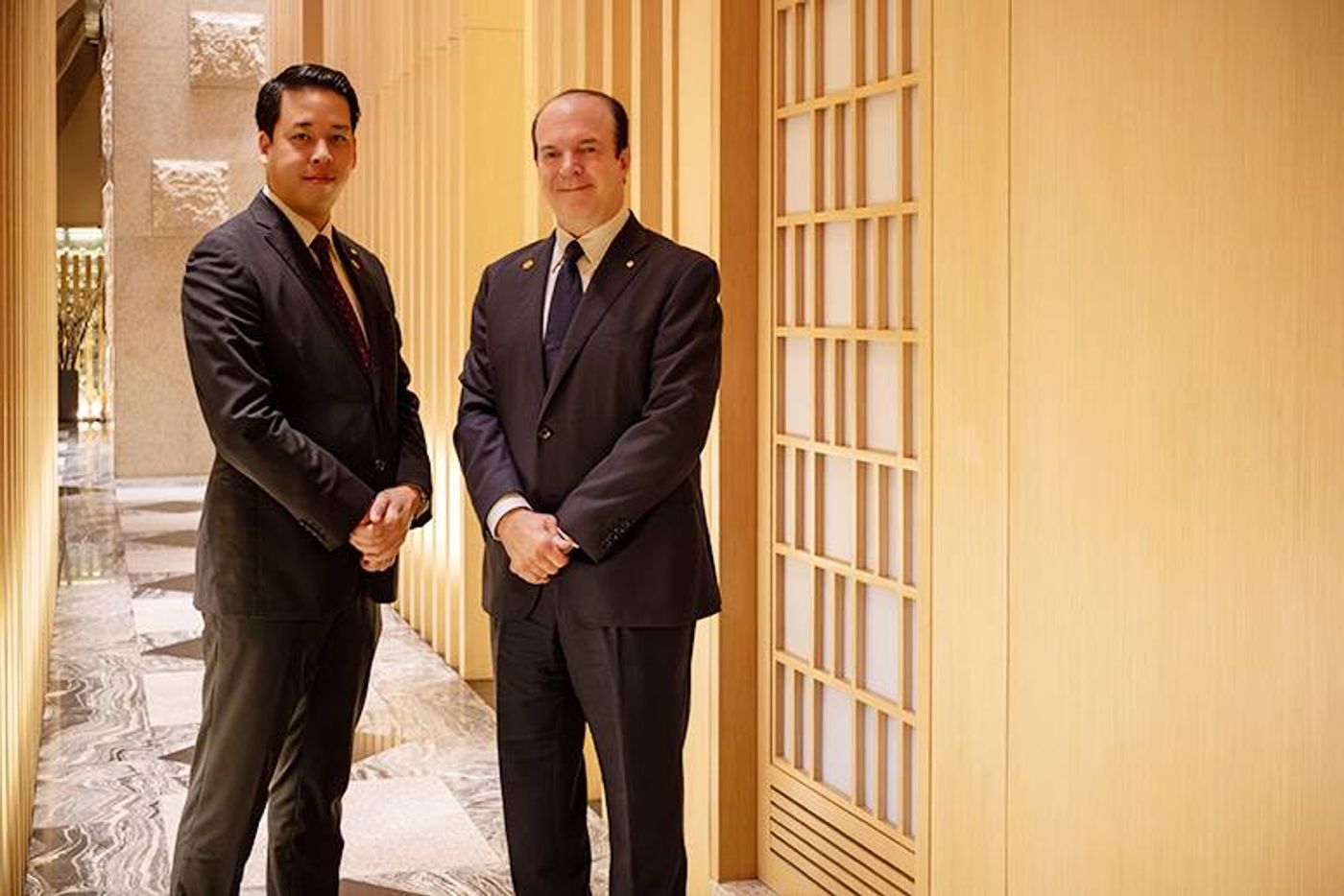 Yoki Takeuchi and Jan William Marshall in dark suits in a wood-latticed corridor of Hotel Okura Manila