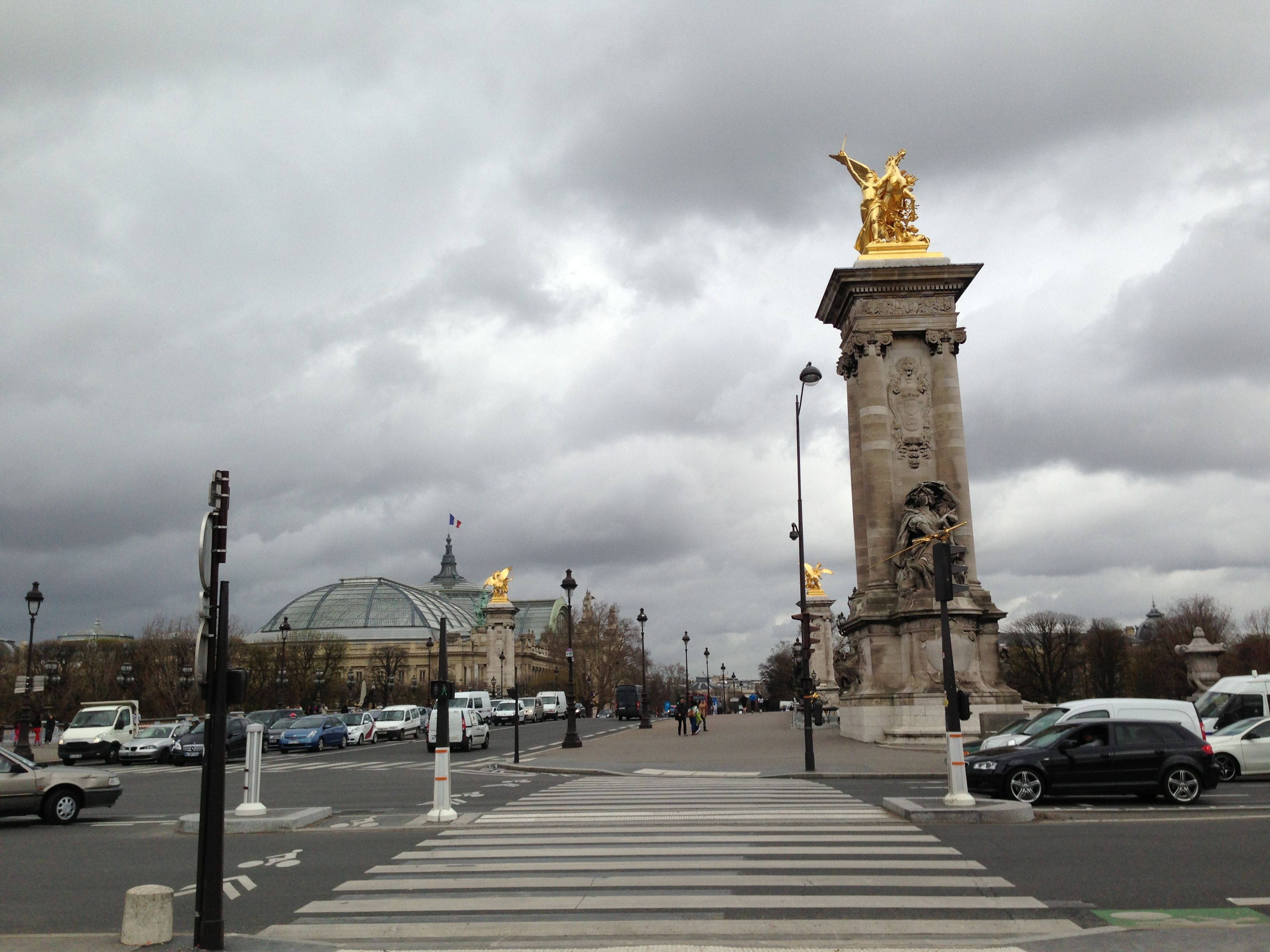 Crosswalk leading toward a stone column topped with a golden winged statue, a glass domed roof beyond