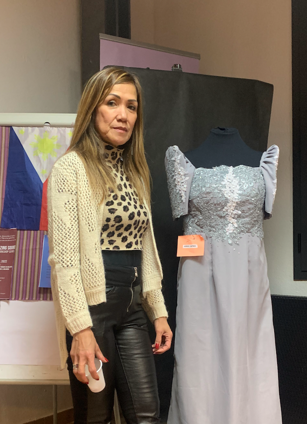 A woman in a cream knit cardigan and leopard-print top stands beside a dress form wearing a silver embroidered Filipiniana gown, a Philippine flag behind her