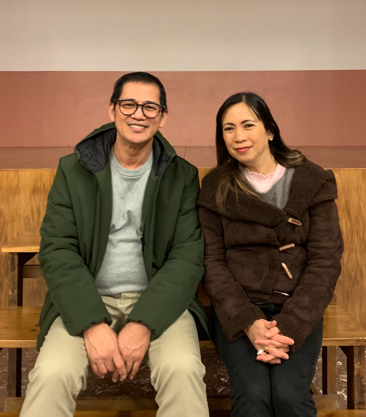 A smiling couple sit side by side on a wooden bench, the man in glasses and a green hooded jacket, the woman in a brown fleece coat