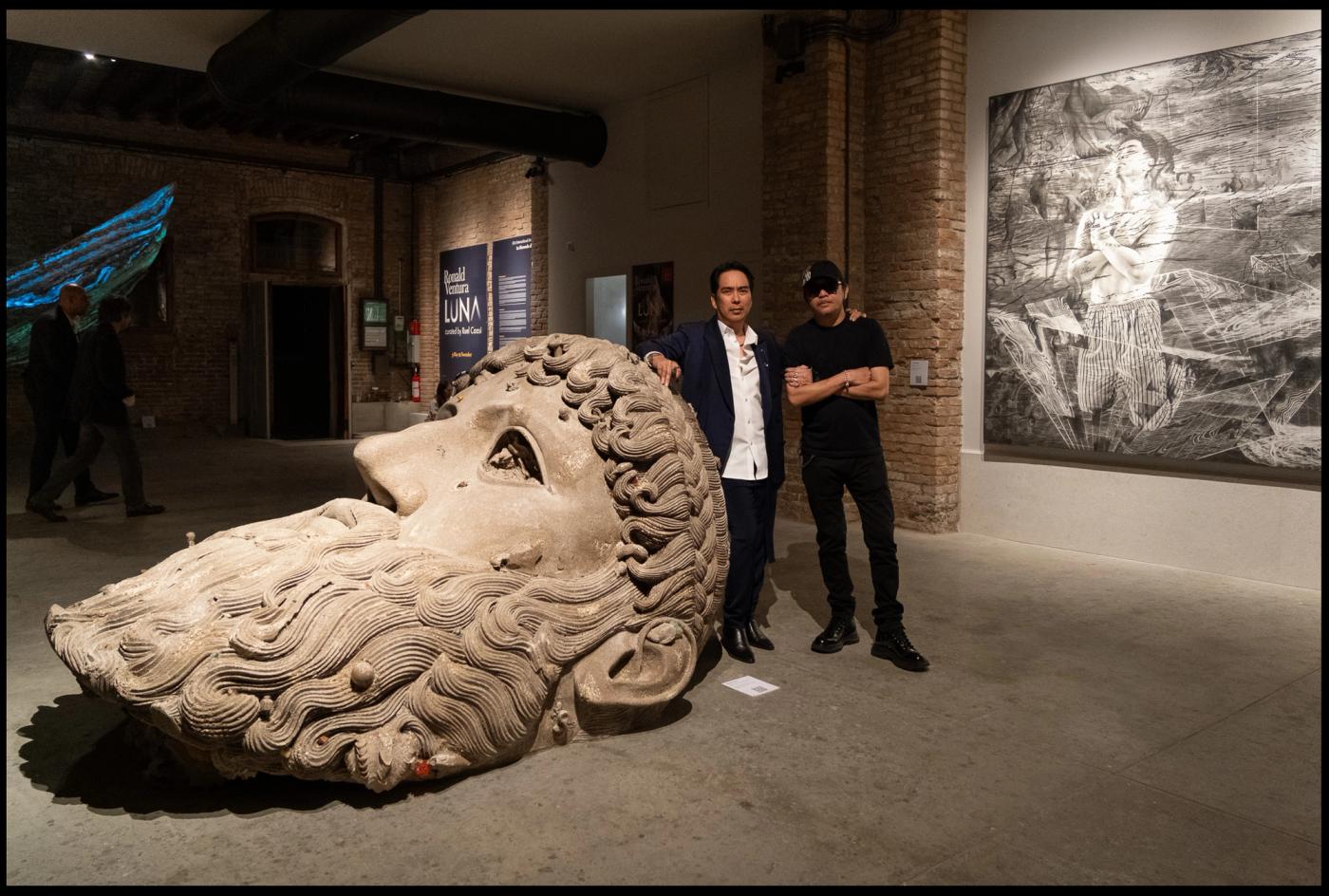 Ronald Ventura in a blue suit and Ruel Caasi in black beside the giant reclining sculpted head of “Luna” in the brick-walled dock hall