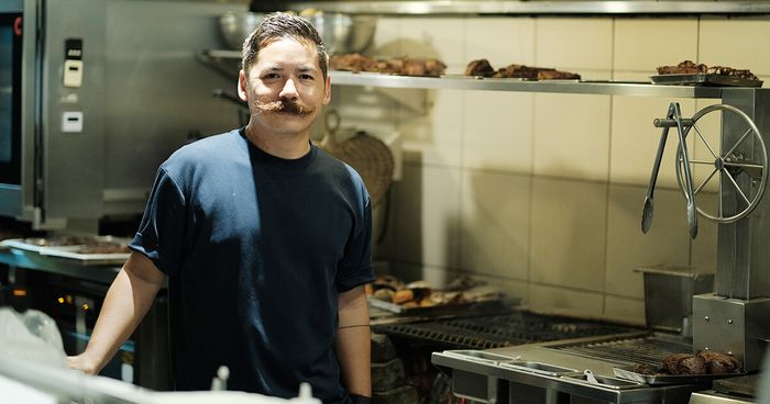 Chef Josh Boutwood with a curled mustache stands in Ember’s kitchen between grills of resting meats