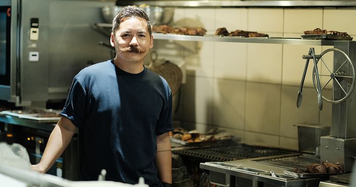 Chef Josh Boutwood with a curled mustache stands in Ember’s kitchen between grills of resting meats