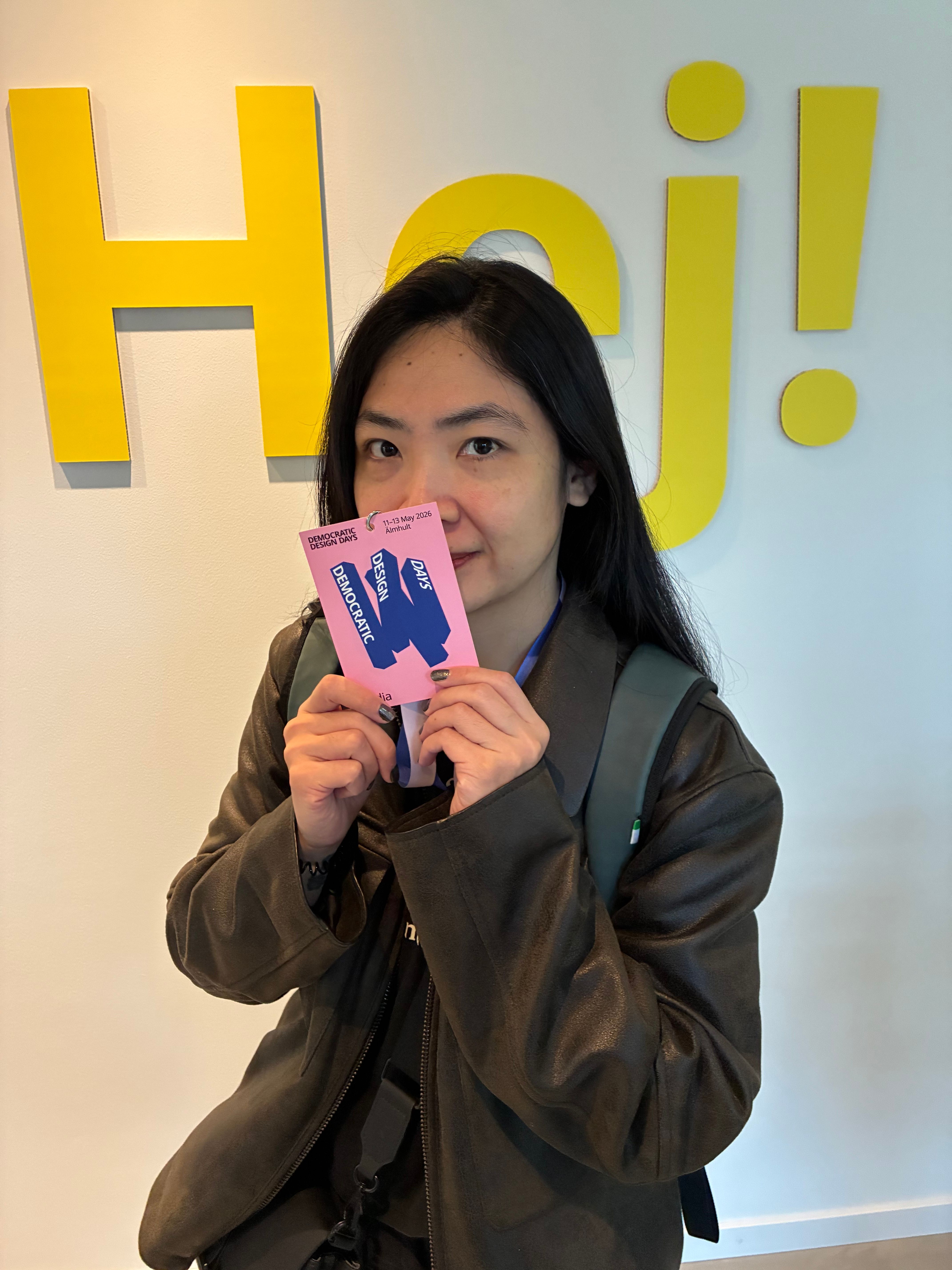 Visitor holding a pink Democratic Design Days badge dated 11-13 May 2026 in front of a yellow Hej! sign in Älmhult
