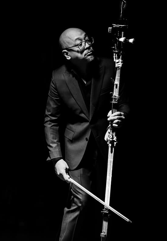 Tad Ermitaño playing the monochord Tungkod staff with a violin bow on stage under dramatic lighting