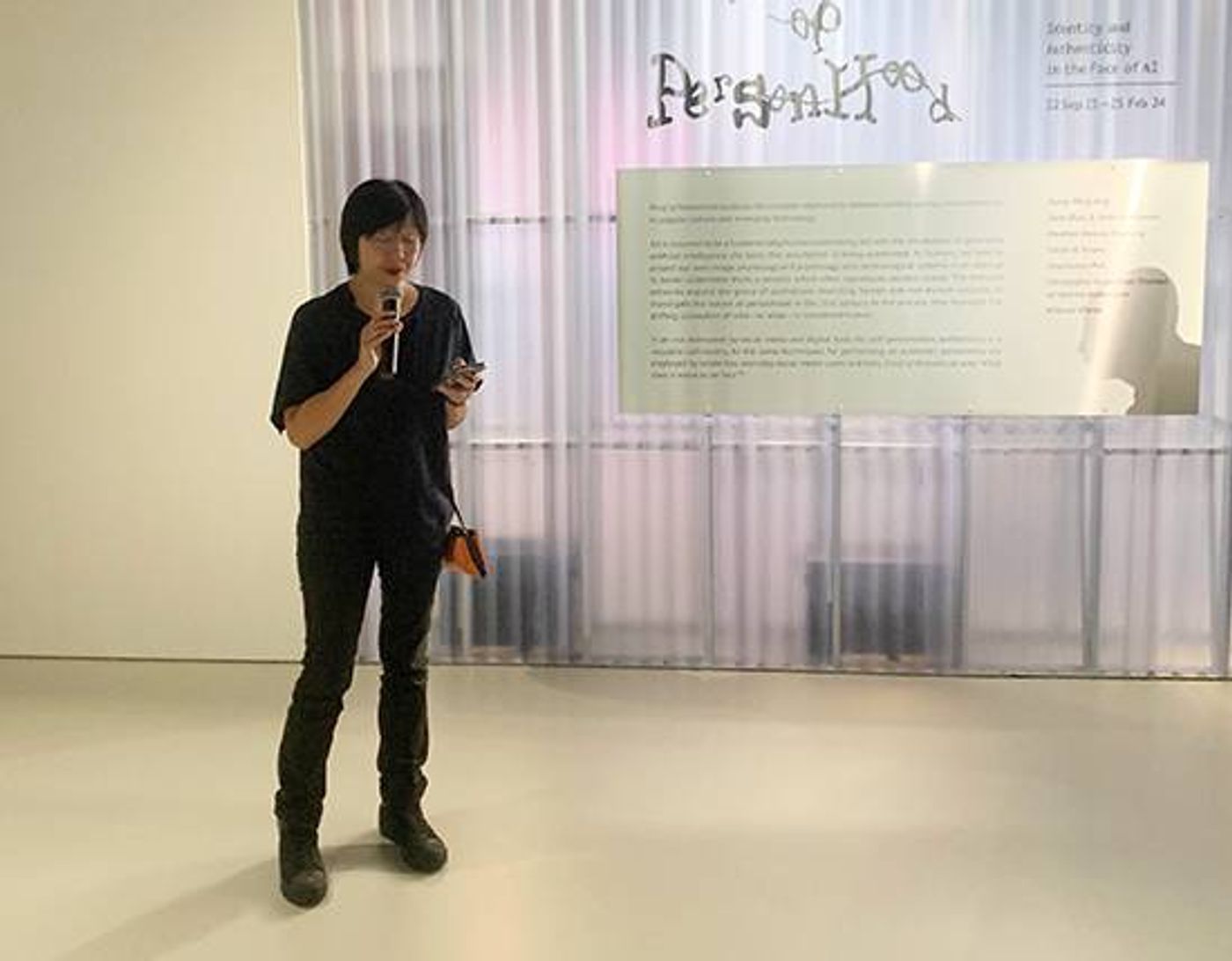 SAM curator June Yap speaking into a microphone in front of the Proof of Personhood exhibition wall text