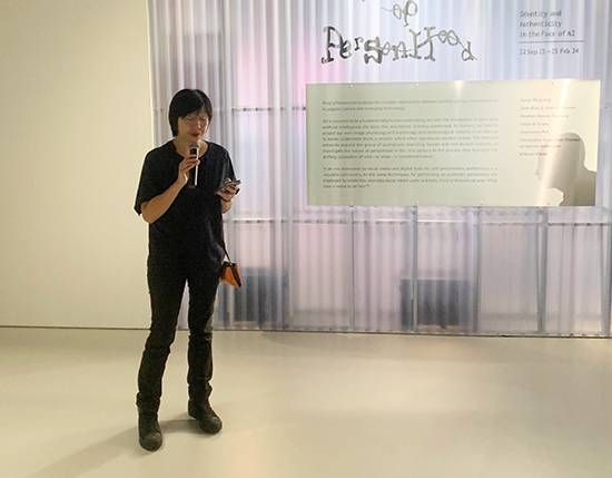 SAM curator June Yap speaking into a microphone in front of the Proof of Personhood exhibition wall text