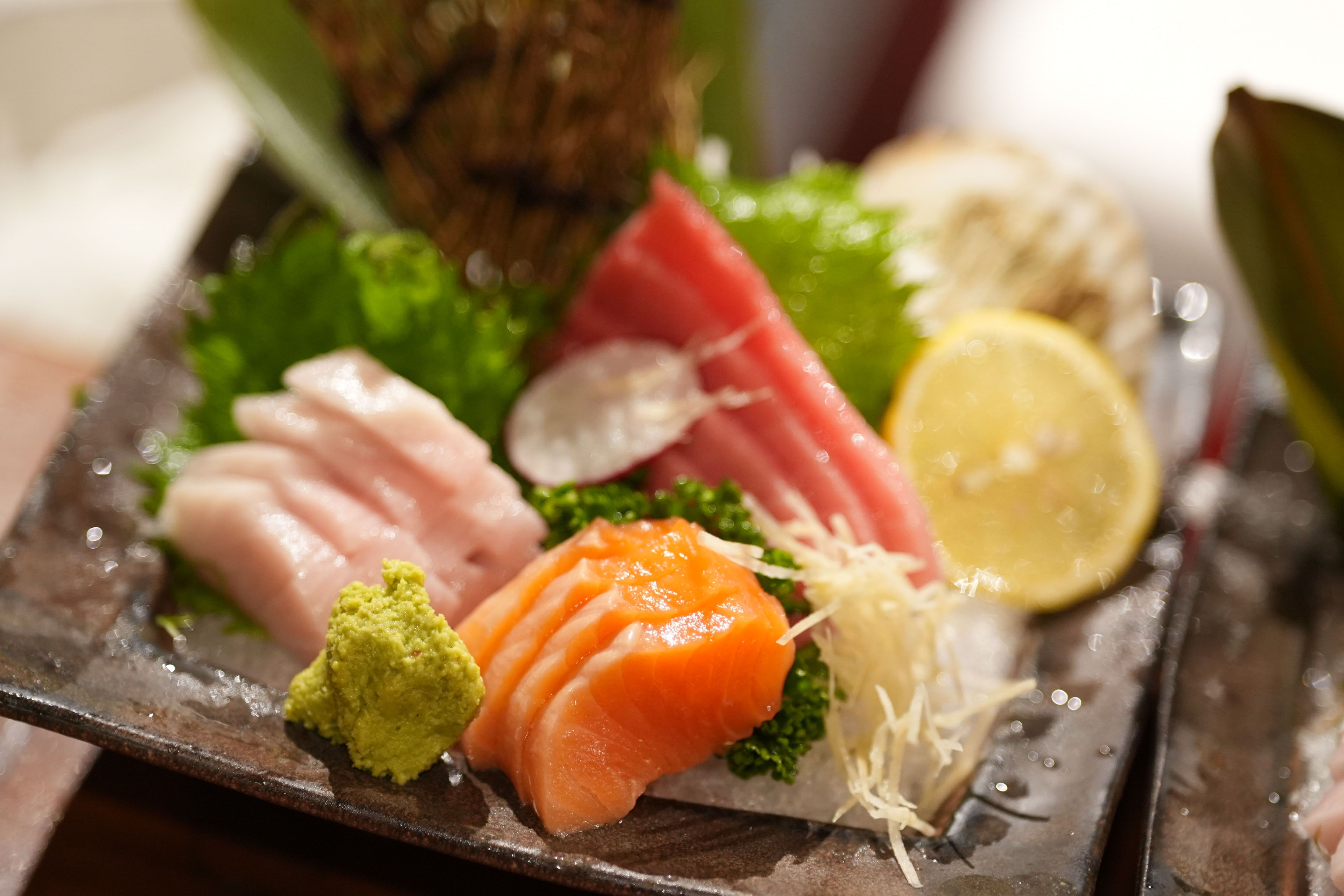 A sashimi platter of salmon, tuna and white fish with wasabi, shredded daikon, shiso and a lemon slice on a dark stone plate