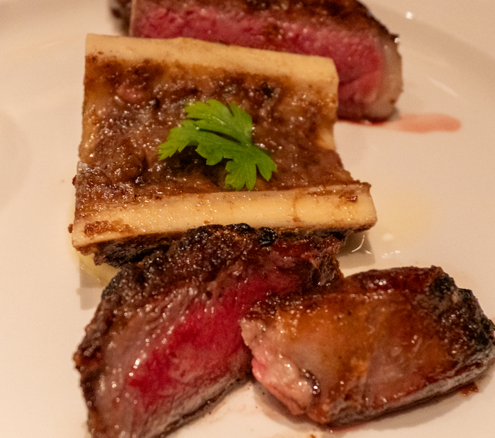 Slices of pink-centered steak beside a roasted split bone marrow garnished with parsley on a white plate
