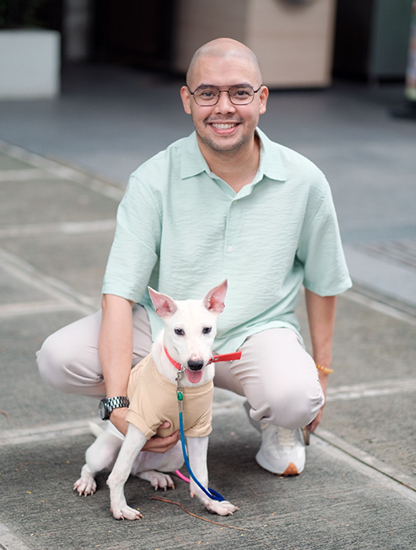 Brian Peck, bald with glasses in a mint polo, crouches beside Shang, a white dog in a beige shirt on a leash