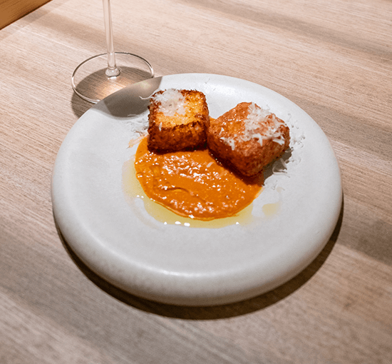Juniper’s tapioca fritters, two golden cubes showered with shaved cheese over a pool of orange sauce