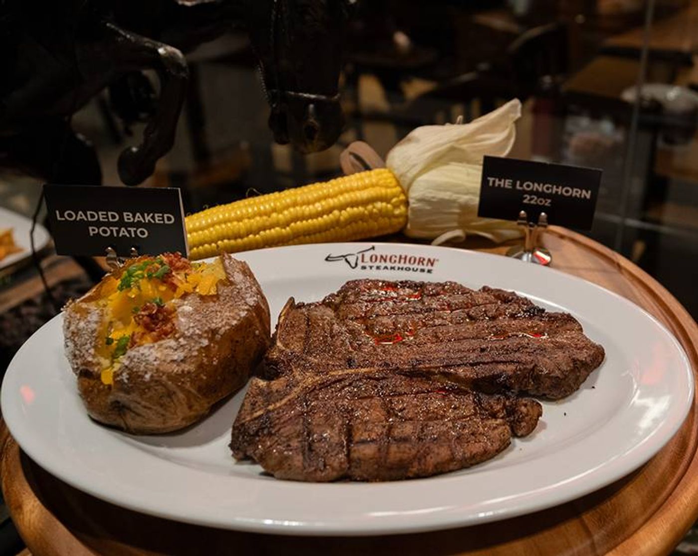 LongHorn porterhouse and a loaded baked potato on a white platter, corn on the cob behind