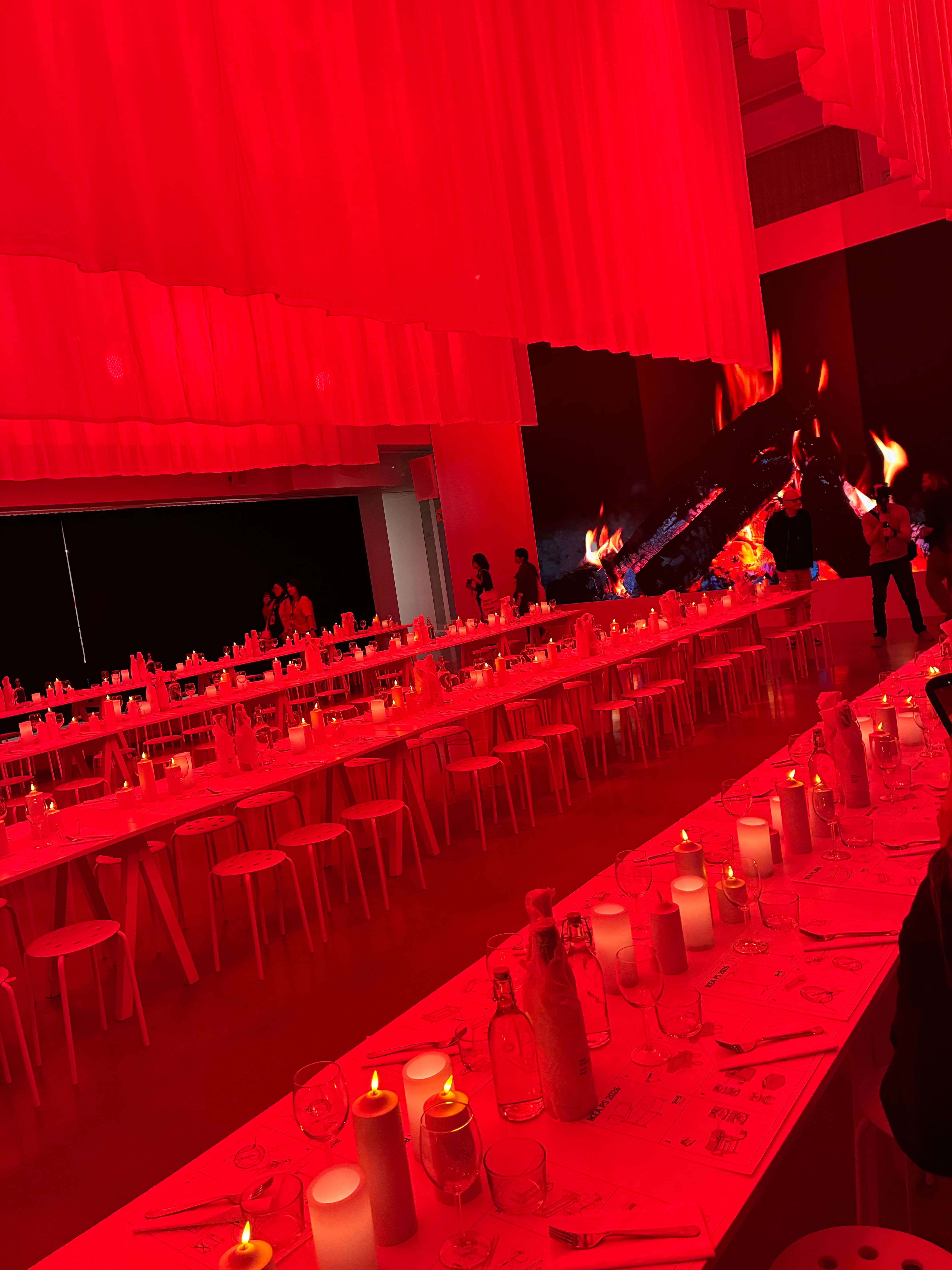 Long candlelit dinner tables under red-lit drapes at a Democratic Design Days banquet, a giant bonfire screen glowing at the far end