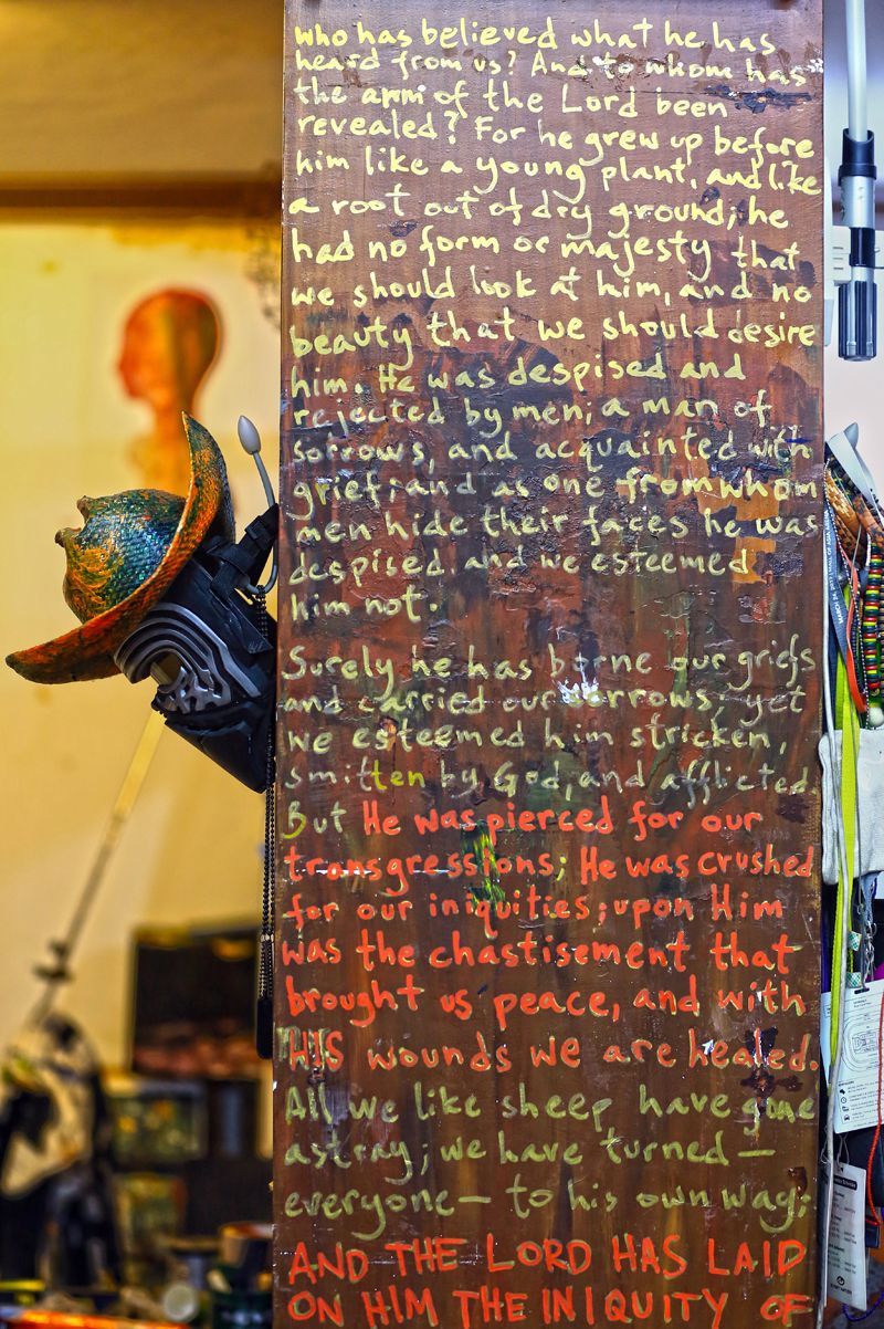 A tall wooden panel covered in hand-lettered text in yellow, orange and green paint, with a painted mask and a helmet hanging on the wall beside it