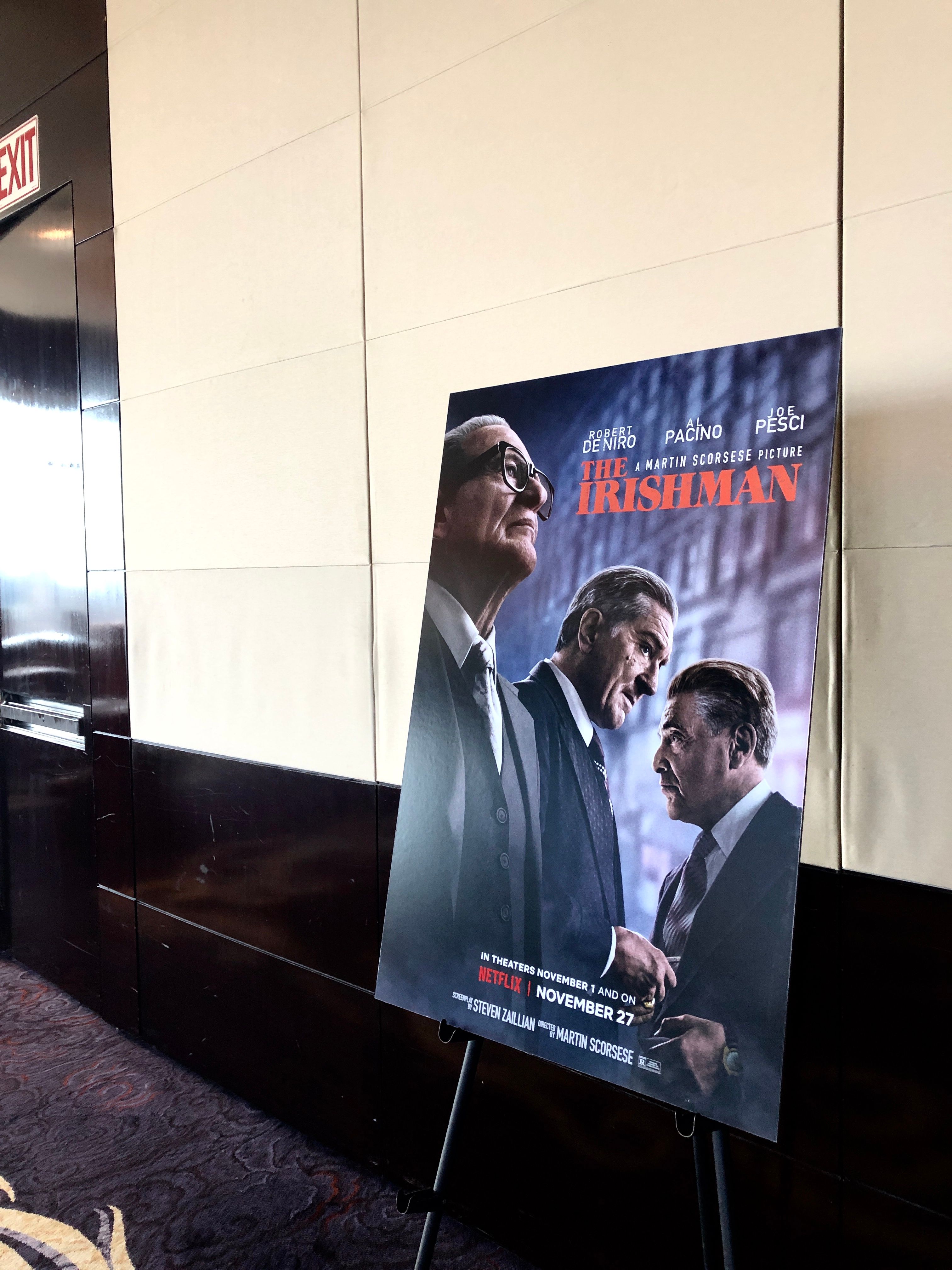 Poster for The Irishman on an easel stand in a dim cinema hallway, billing De Niro, Pacino and Pesci