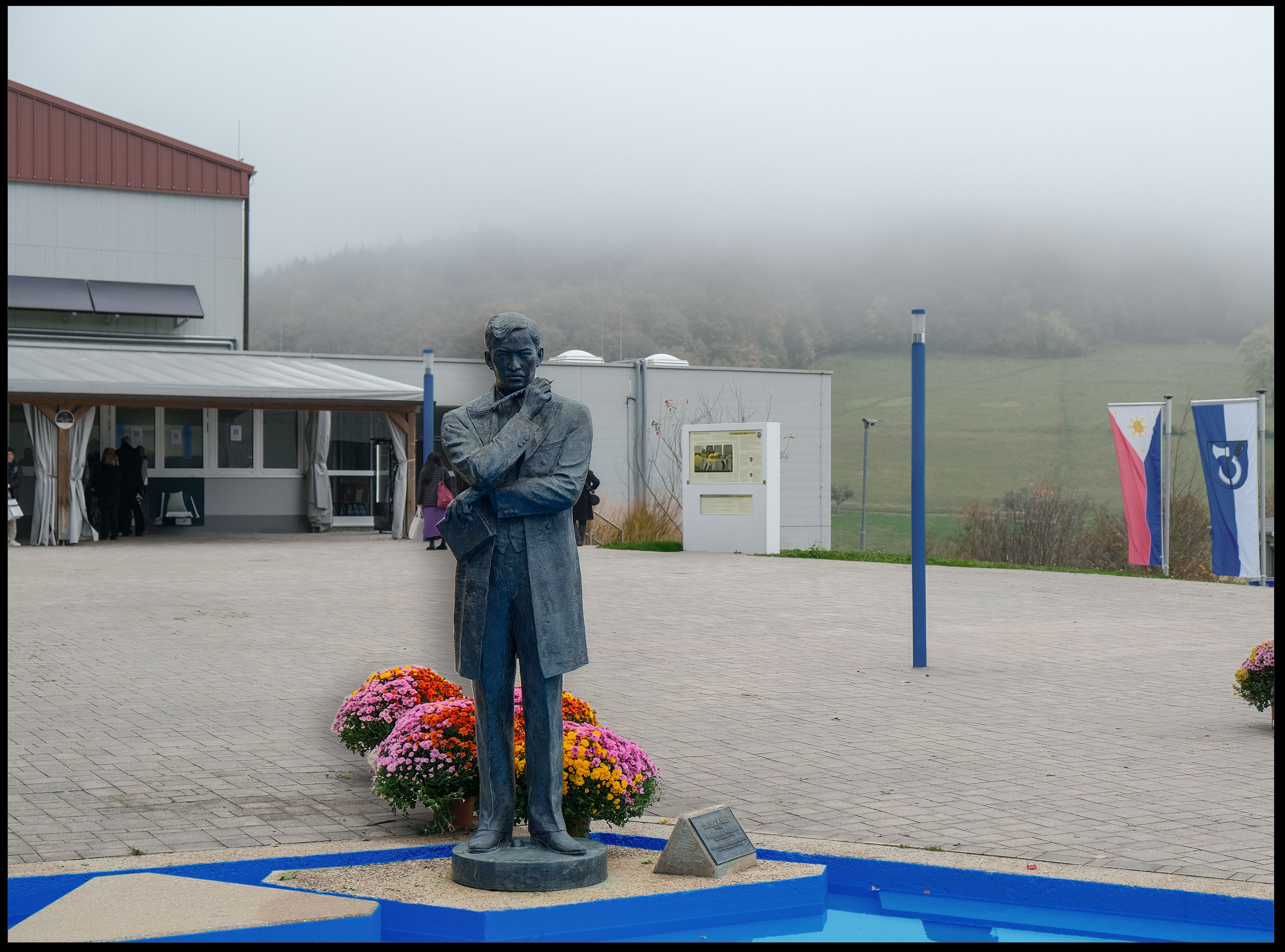 Bronze statue of José Rizal in an overcoat, chin resting on his hand, with bright chrysanthemums at its base and a Philippine flag over the misty hills behind