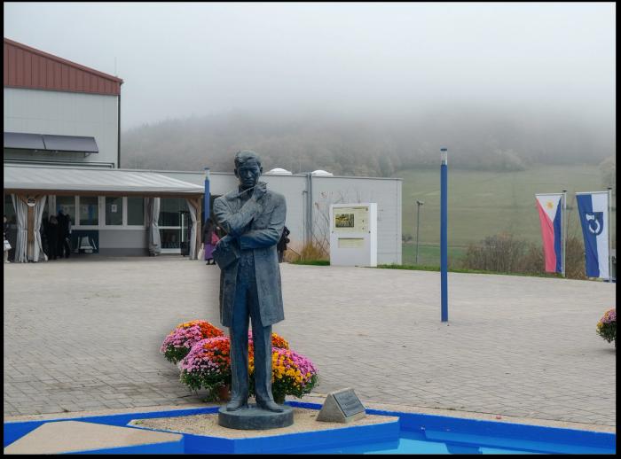 Bronze statue of José Rizal in an overcoat, chin resting on his hand, with bright chrysanthemums at its base and a Philippine flag over the misty hills behind