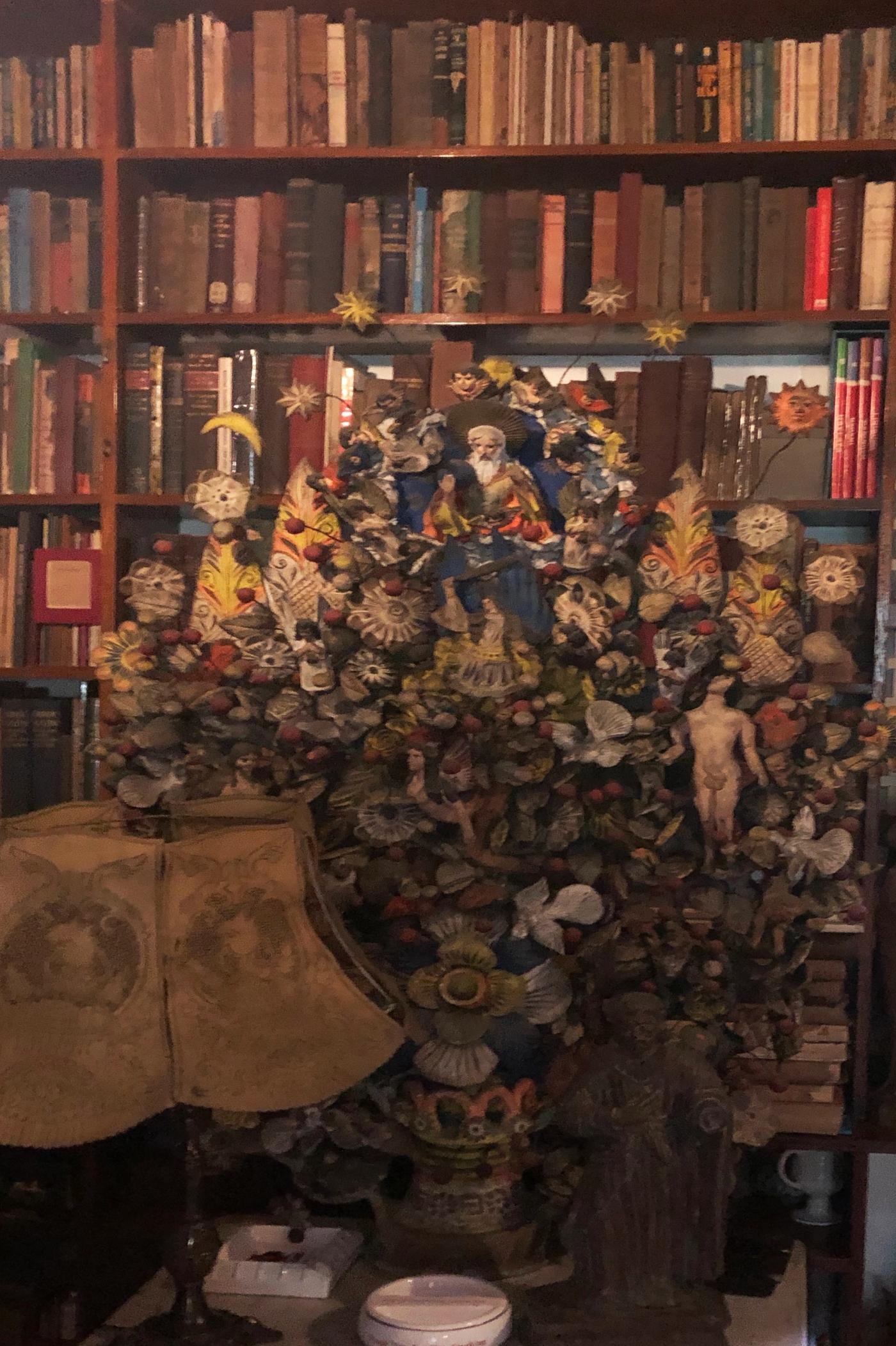 A polychrome clay tree of life crowded with flowers, birds, shells and small figures, standing in front of crowded bookshelves